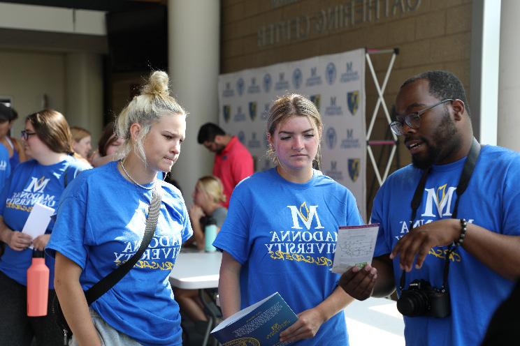 Three students wearing Madonna University t-shirts at orientation are listening to each other speak 和 looking at a piece of paper intently